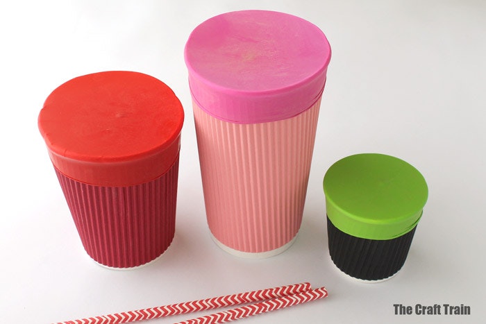 3 different sized paper cups with balloons stretched over top of all three to create paper cup drums. With paper straws next to them for drum sticks
