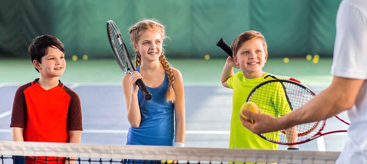 3 kids with tennis rackets smiling waiting to learn how to play tennis