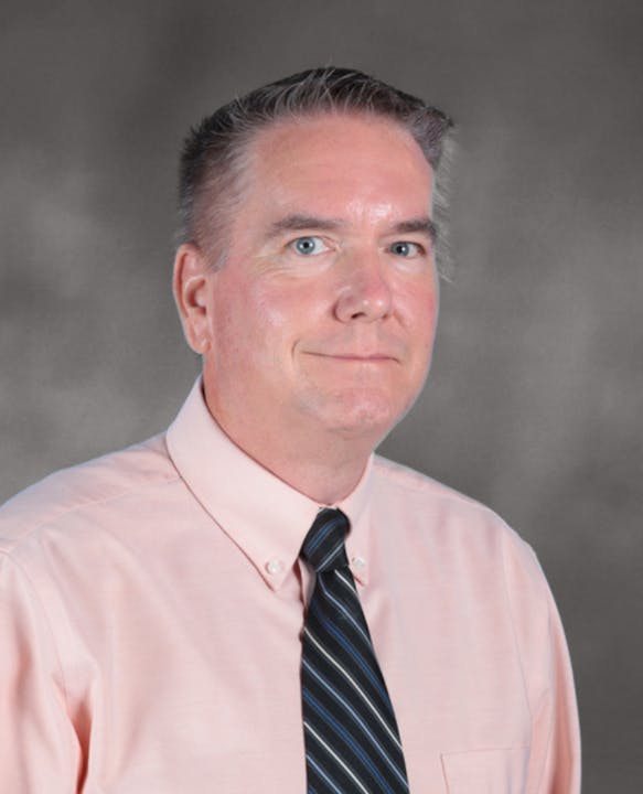 A person wearing a light pink shirt and a striped tie, against a gray background.