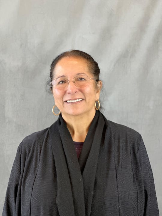 A smiling woman with earrings, glasses, and a black shawl against a textured grey backdrop.