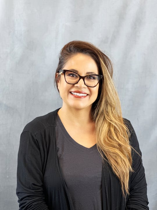 A smiling person with glasses, black clothing, against a grey backdrop.