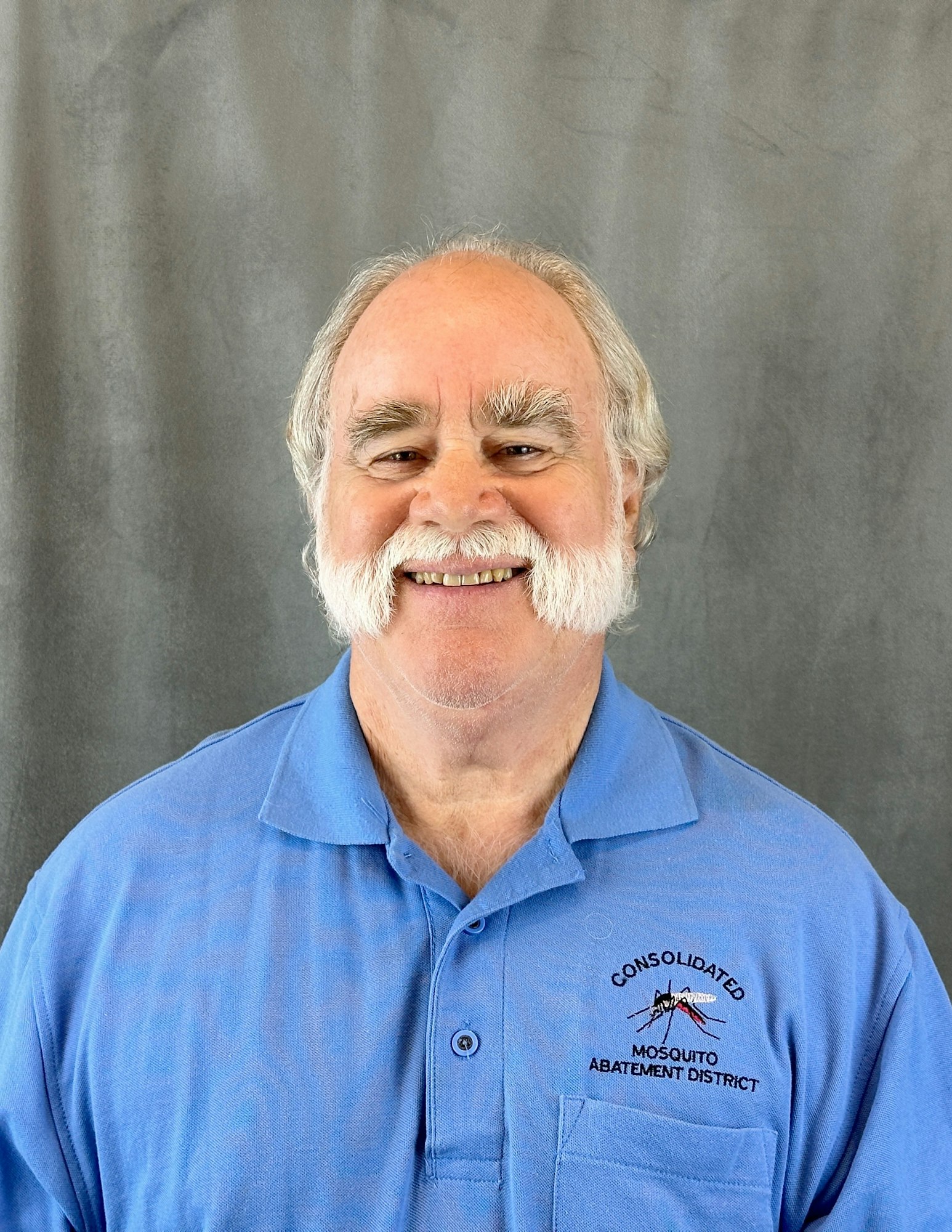A smiling man in a blue shirt with an embroidered logo: "Consolidated Mosquito Abatement District." Gray background.
