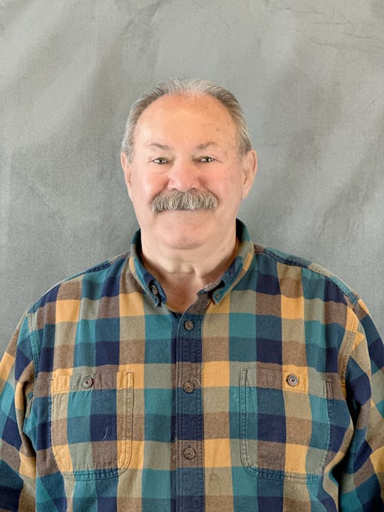 Man with a mustache wearing a plaid shirt, standing against a grey backdrop.