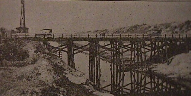 A vintage photo of a wooden bridge over a river with a car and a tower in the background.