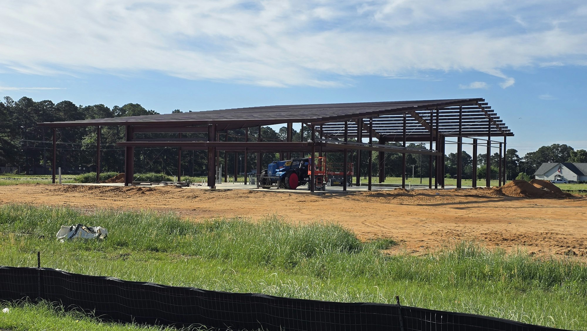 The image shows a construction site with a metal building framework, heavy machinery, and a grassy area.