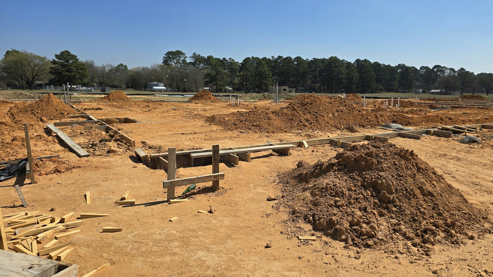 A construction site featuring earth moving and wooden forms, with piles of dirt and framing materials under a clear blue sky.