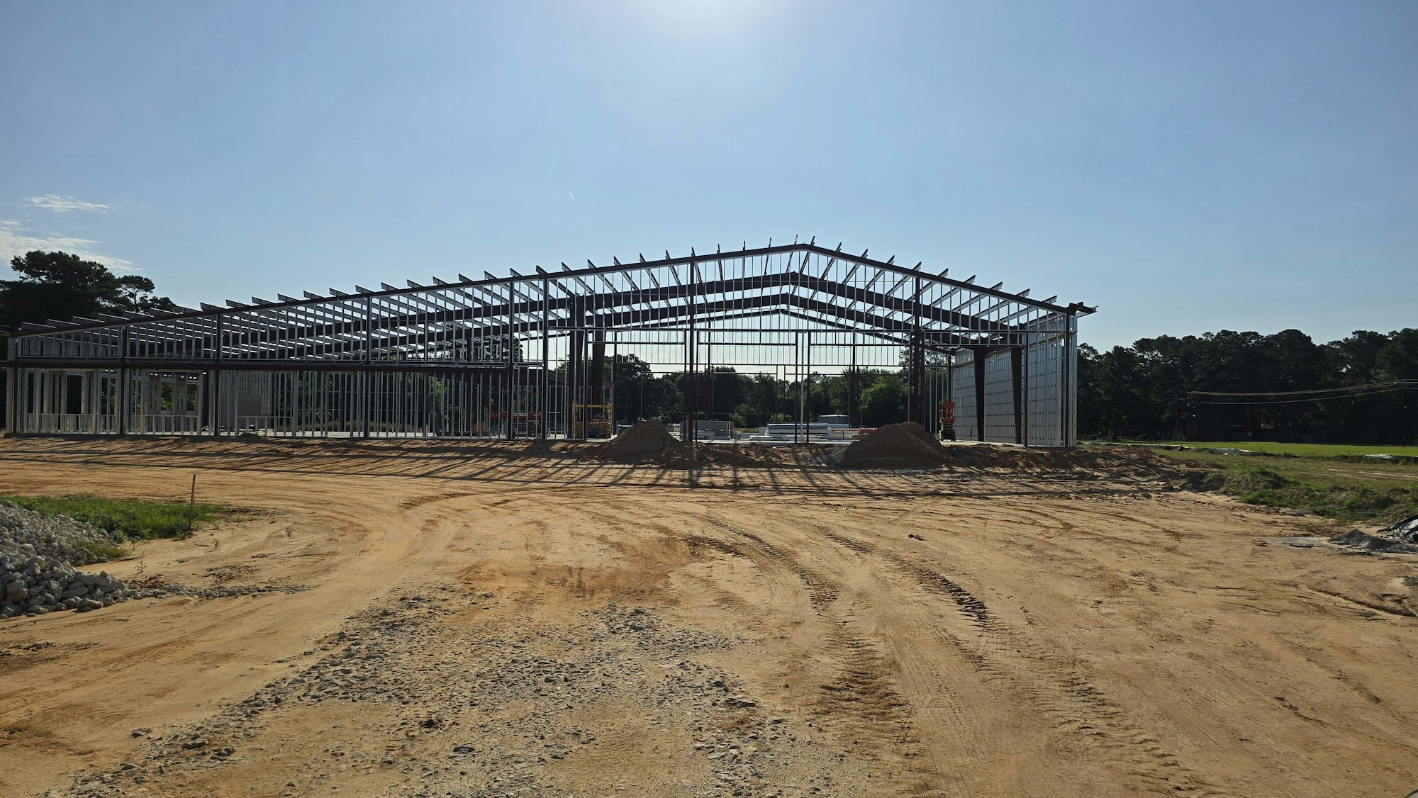The image shows an under-construction building with metal framing against a clear sky, surrounded by dirt and gravel.