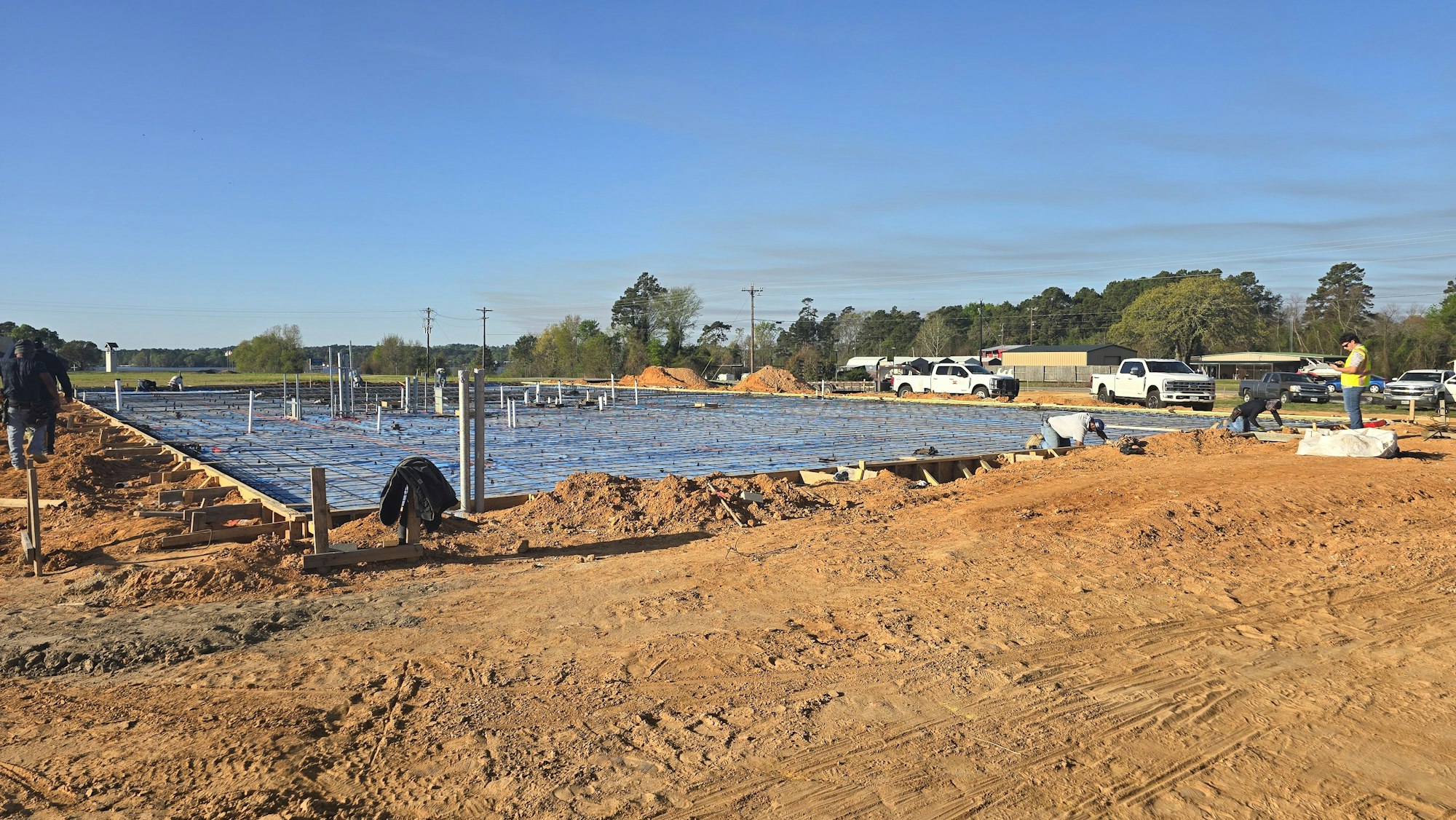 A construction site with workers preparing a concrete slab, surrounded by machinery and clear blue skies.