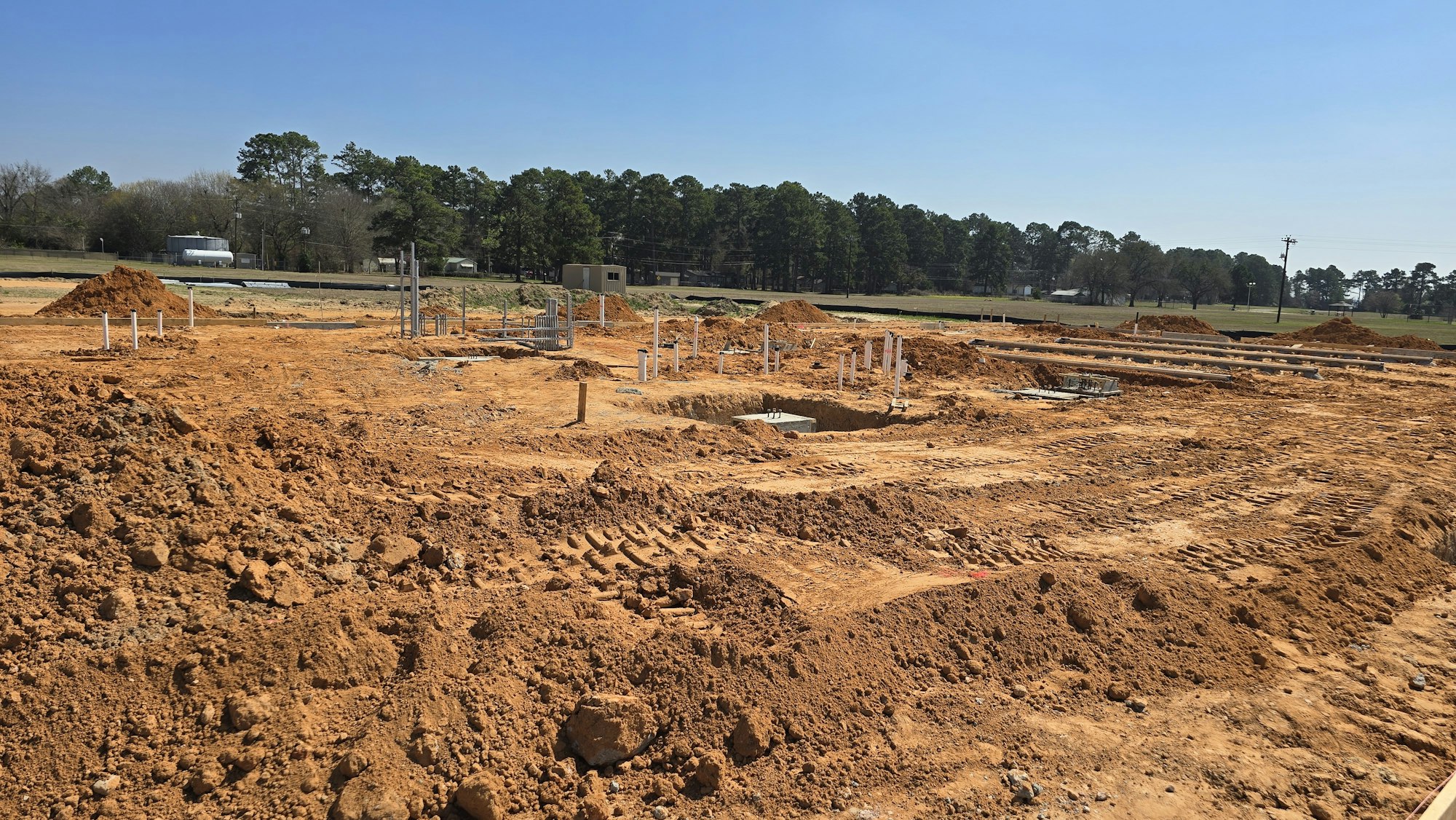 A construction site with dirt, piles of earth, and foundation work in progress under a clear blue sky.