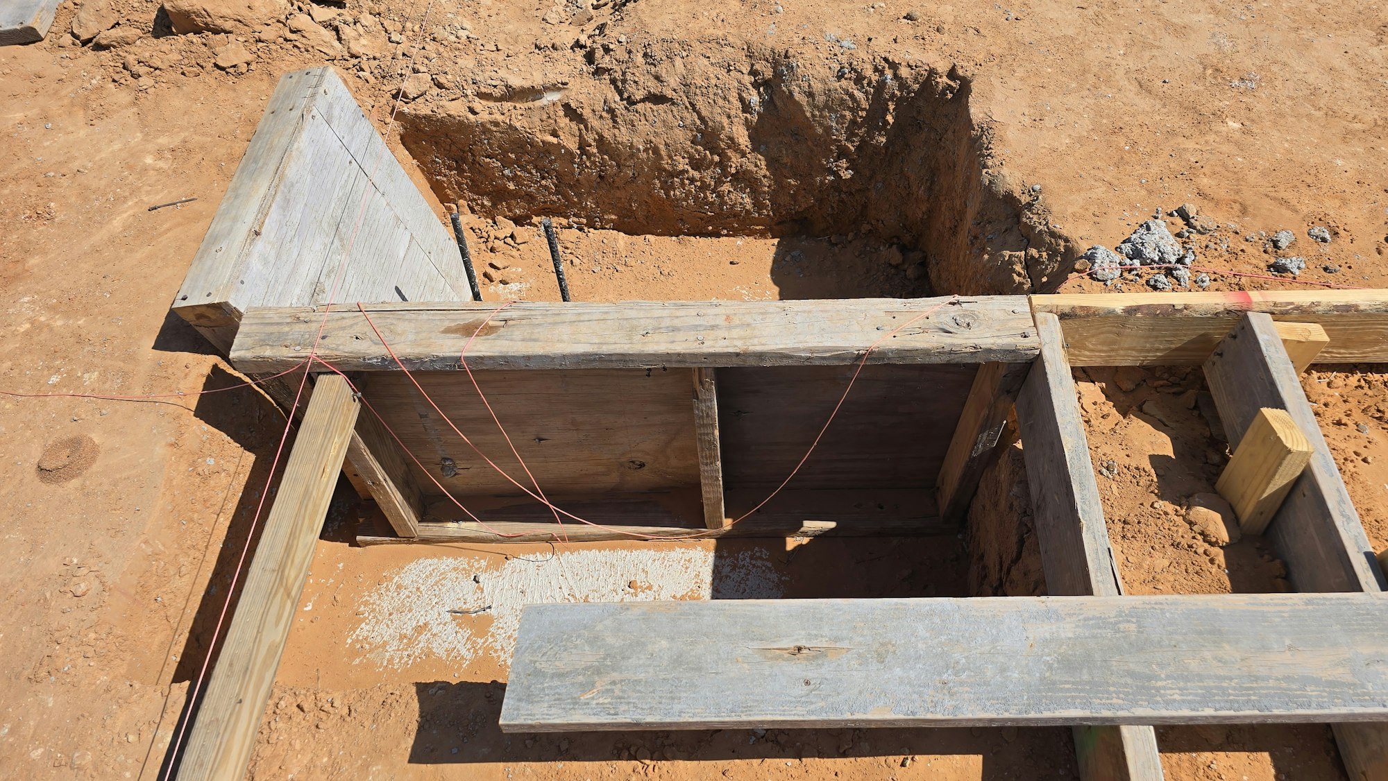 The image shows a construction site with wooden framework and a deep excavation in sandy soil.