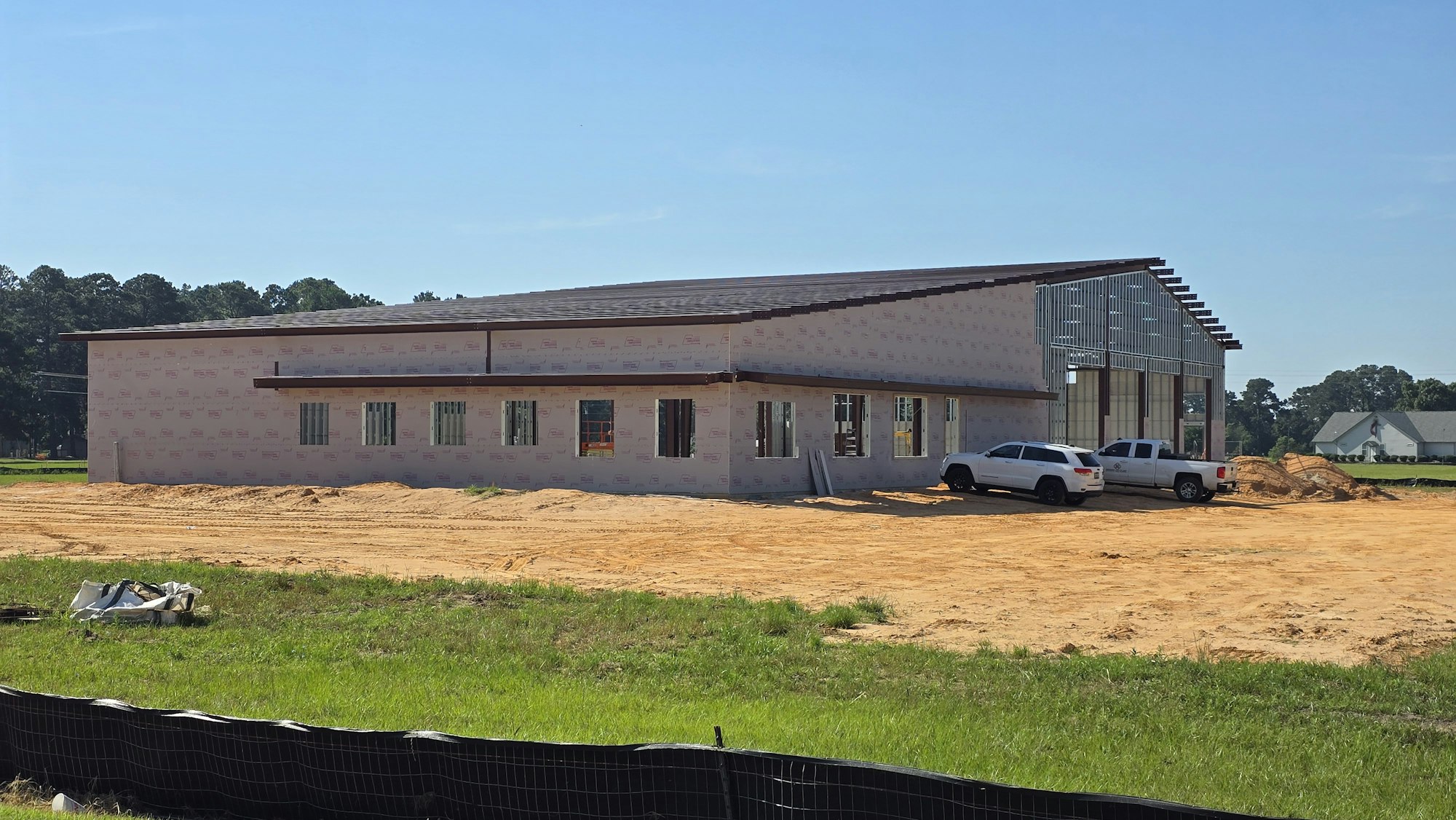 A partially constructed building on a sandy lot with two white trucks parked nearby, under a clear blue sky.