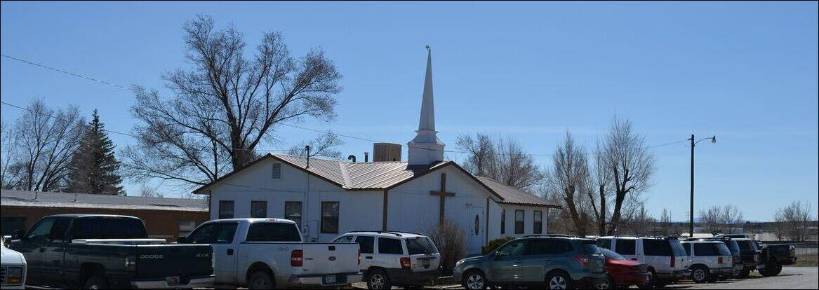 A small white church with a steeple, several parked cars, and bare trees in the background under a clear blue sky.