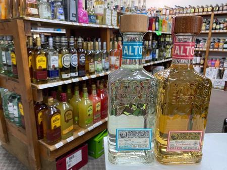 Two bottles of tequila on display in a liquor store with various other alcohol bottles on shelves in the background.
