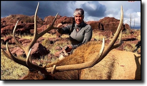 A person outdoors with a large deer, likely an elk, in a rocky and grassy landscape under a cloudy sky.