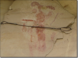 Ancient rock art depicting a humanoid figure with raised arms and faded red pigment on a stone surface, partially obscured by a dark cable.