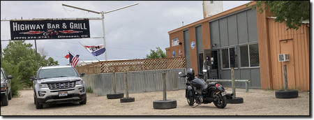 A bar and grill with a sign, parked car, motorcycle, and flags.