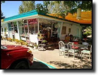Outdoor seating area of a retro diner with a red car parked outside. Sign reads "Malts" and "Shakes." Tables, chairs, and plants visible.