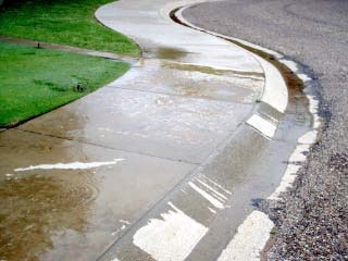 Curving sidewalk with puddles after rain, next to a grassy area and gravel.