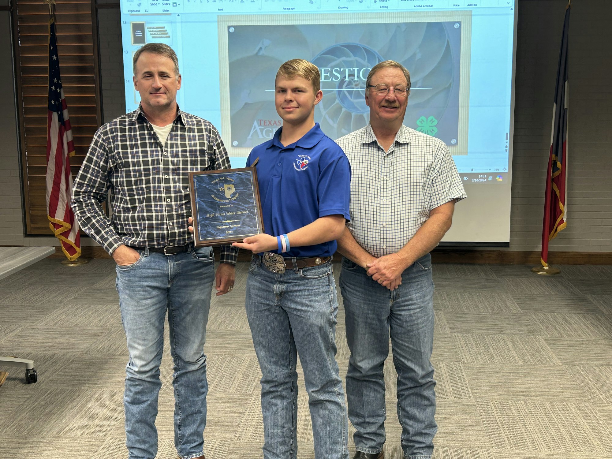 Three people posing with an award plaque. US flag and Texas state flag in the background, with a presentation screen.