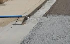 Water flows from a hose onto a curb beside wet asphalt, likely indicating street cleaning or maintenance.