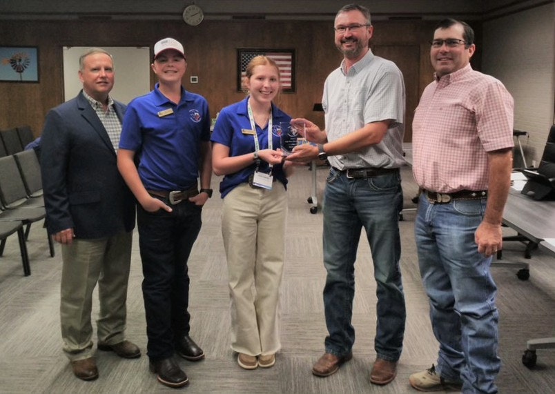 Five people standing indoors; two in blue shirts holding an award, smiling.