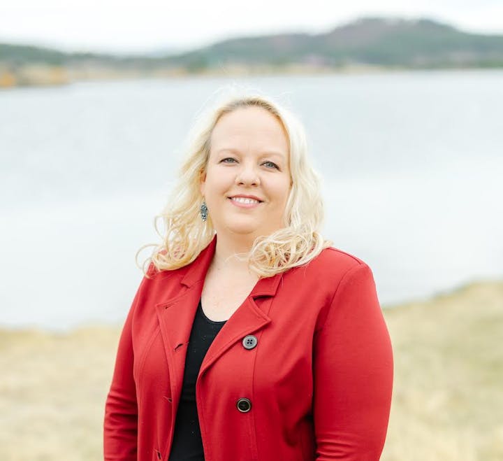 A woman with long blonde hair wearing a red blazer over a black top stands outdoors in front of a calm body of water with hills in the background. She is smiling and facing the camera.