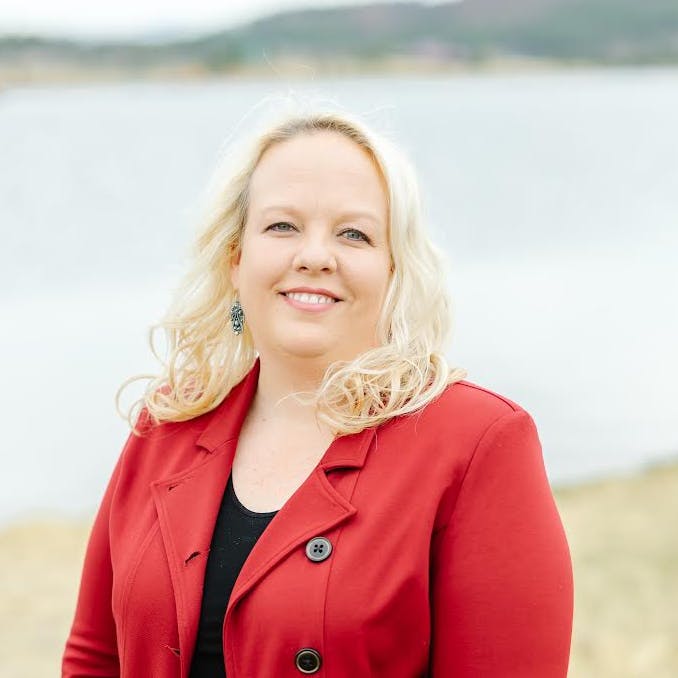 A woman with long blonde hair wearing a red blazer over a black top stands outdoors in front of a calm body of water with hills in the background. She is smiling and facing the camera.