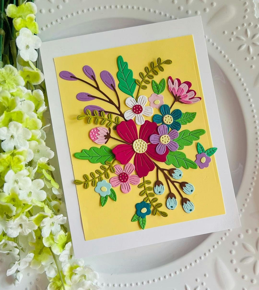 A colorful floral card featuring layered paper flowers and leaves on a yellow background, alongside white decorative flowers.