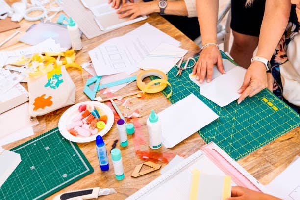 A crafting table with papers, glue, scissors, and art supplies, with people making crafts hands-on.