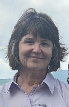 A smiling woman with mid-length hair, wearing a light purple shirt, with a cloudy sky in the background.
