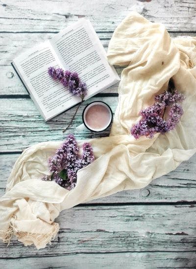 An open book, lilac flowers, a cream scarf, and a jar of cream on a wooden surface.