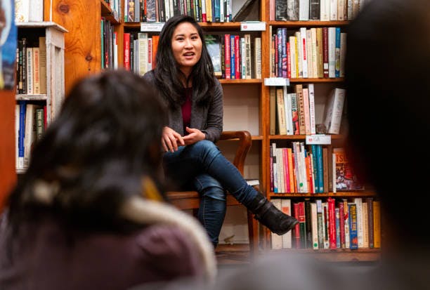 A woman speaking in a bookstore, seated in front of bookshelves, with people listening in the foreground.