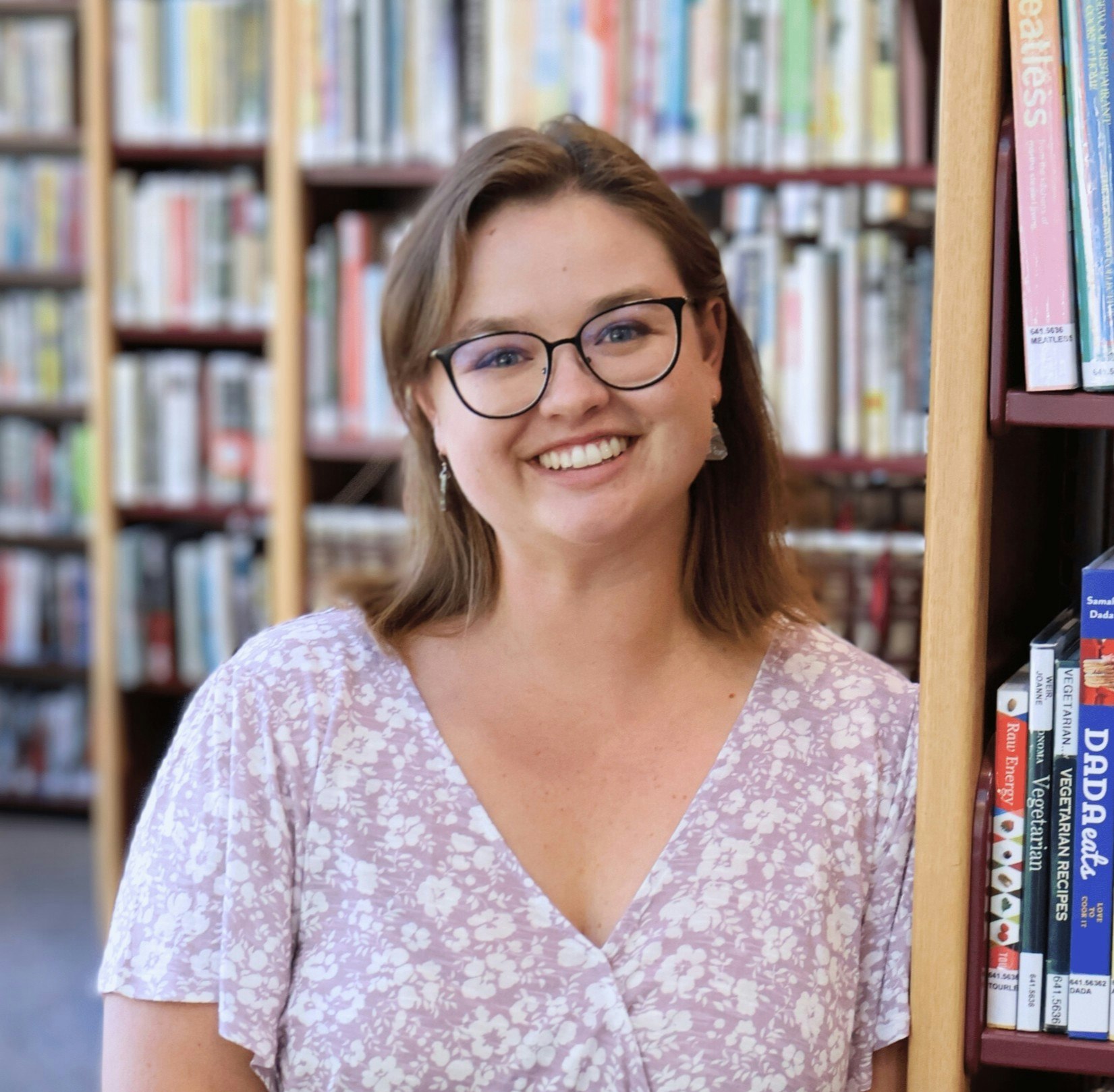 A person in glasses is smiling in front of bookshelves in a library.