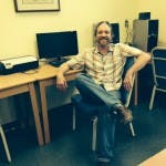 May contain: furniture, table, clothing, pants, desk, person, sitting, computer, electronics, adult, male, man, chair, face, head, photography, portrait, computer hardware, hardware, monitor, and screen