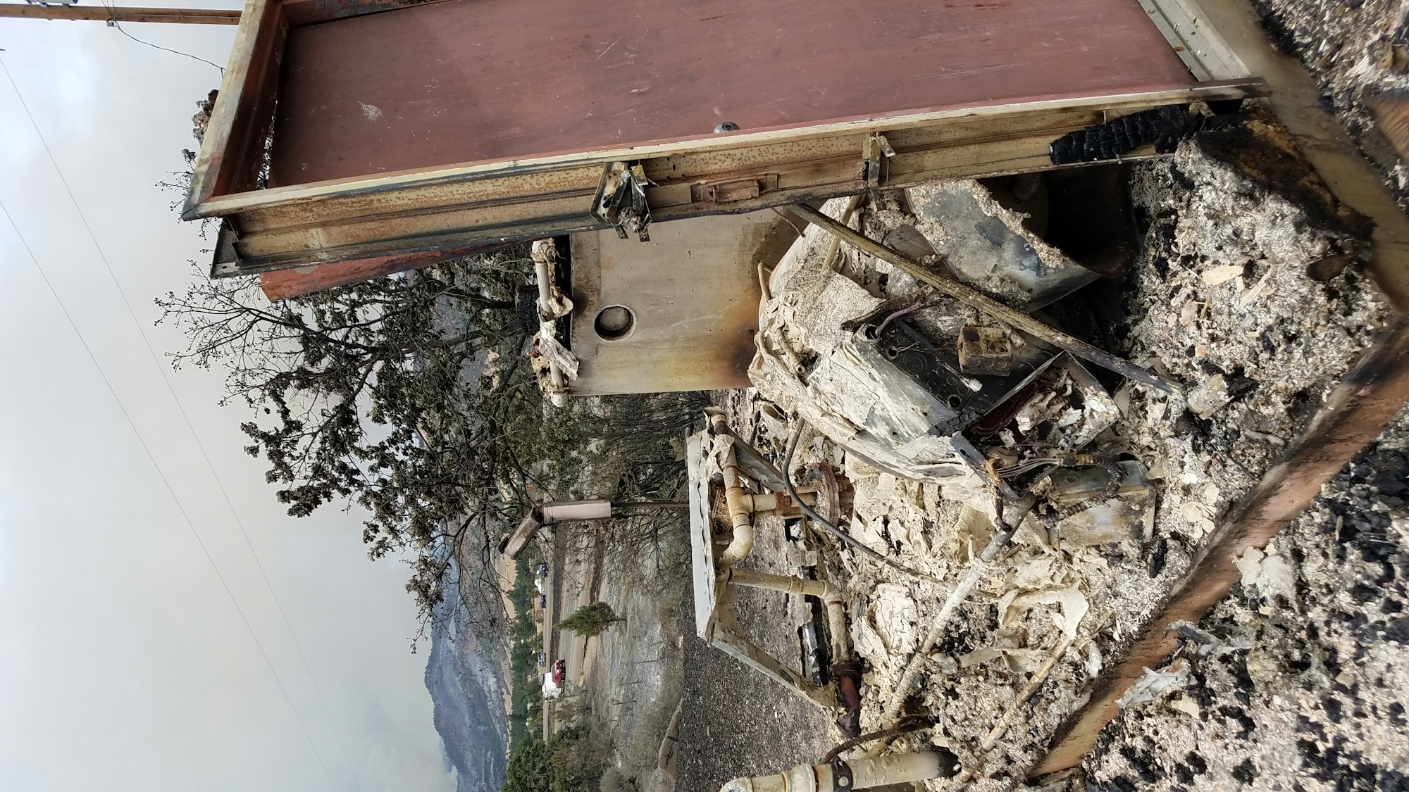 Burned structure in a mountainous area showing extensive fire damage. A metal door stands upright amidst ash, debris, and collapsed building materials. Charred remnants of furniture and framing are visible, with smoke still present in the background.
