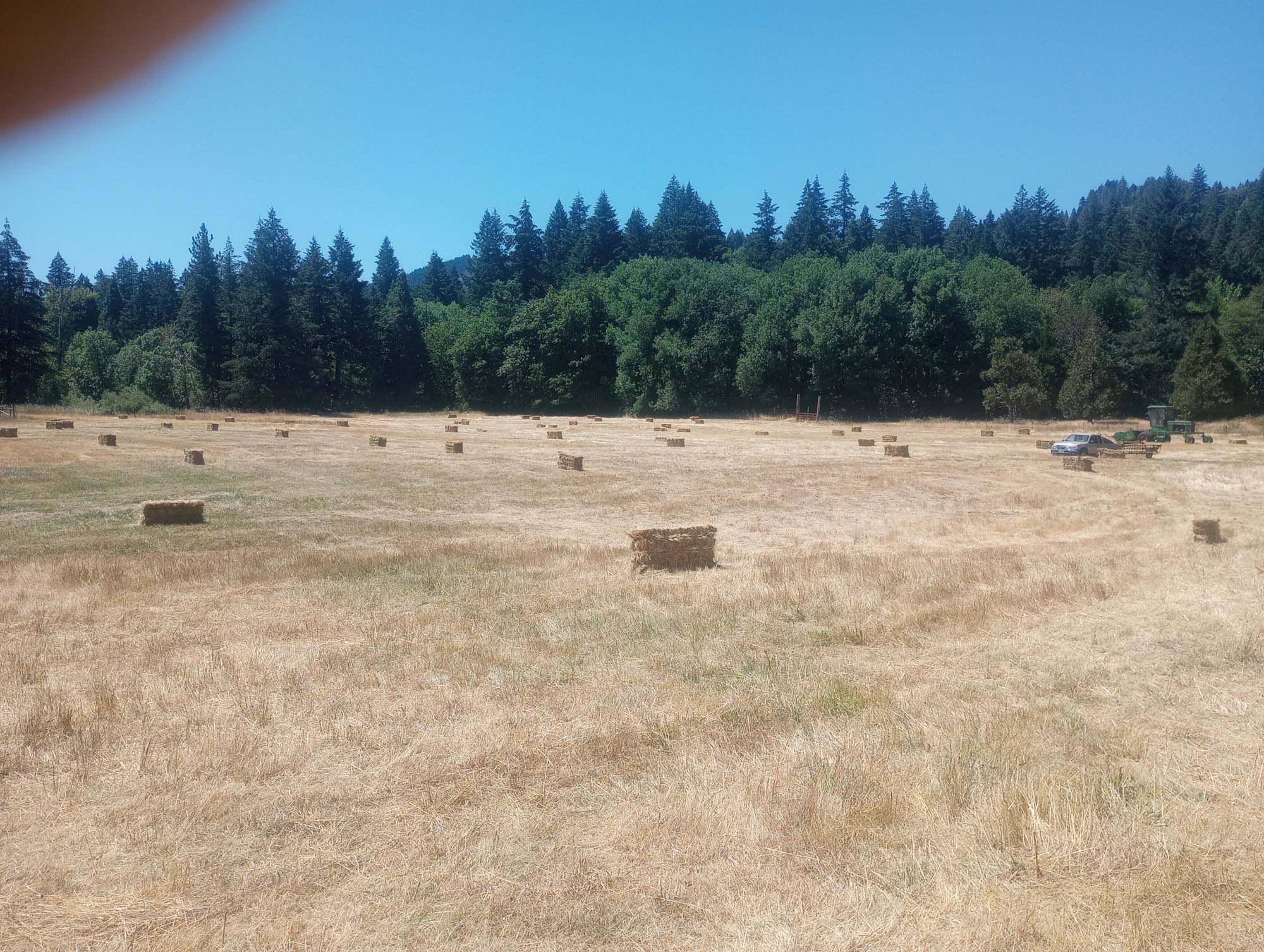 A field with hay bales, trees in the background, and farming equipment. Finger partially obscures the top-left corner.