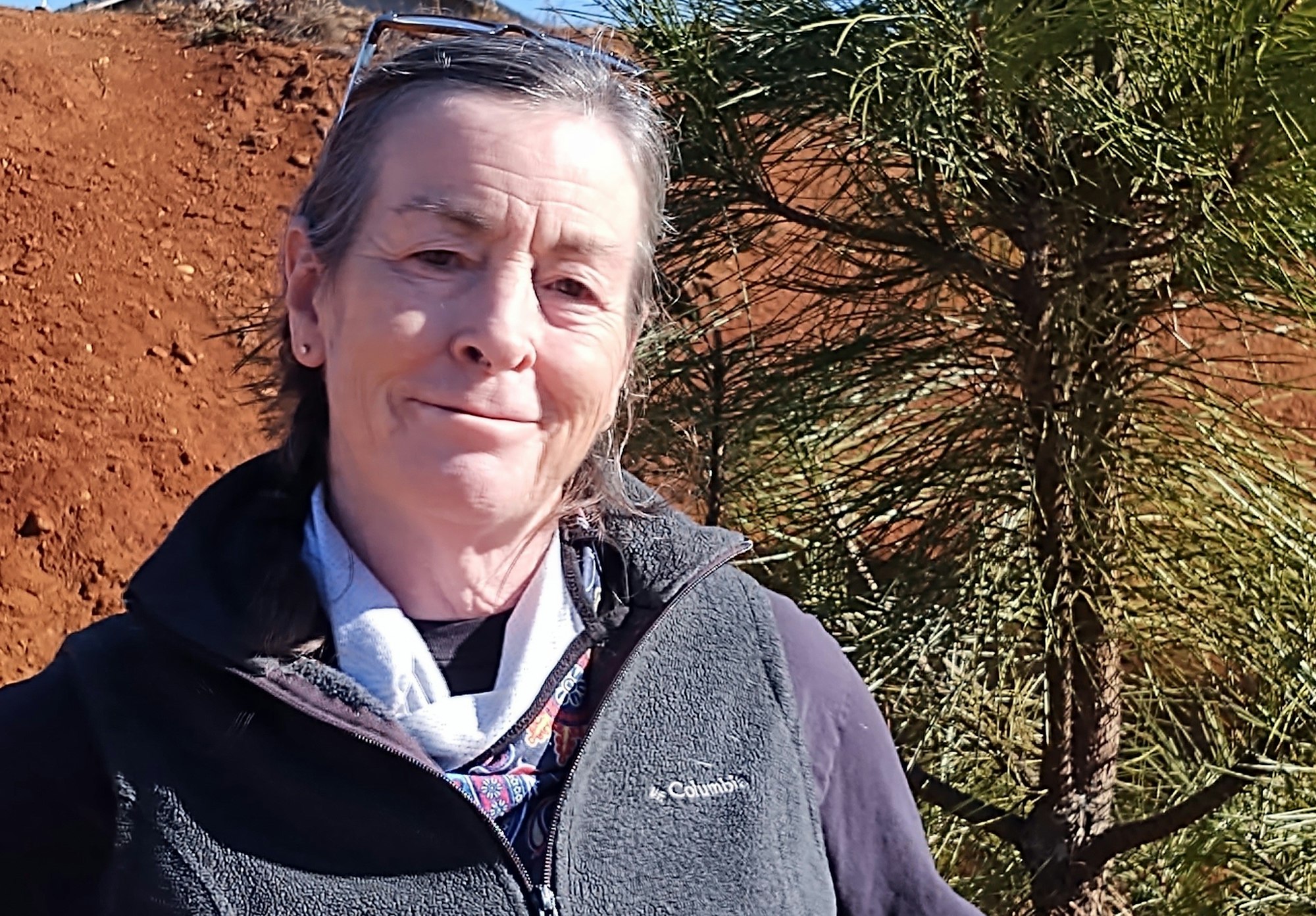 A person with long hair, wearing a vest, smiles outdoors near a tree and reddish soil in a sunny setting.
