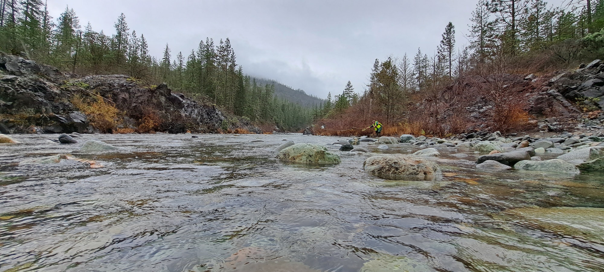 A person in high-visibility clothing stands by a rocky river in a misty forested area.