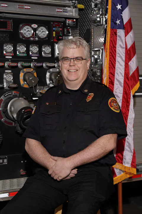 A smiling individual in a firefighter uniform sits in front of a fire truck's control panel and a U.S. flag.
