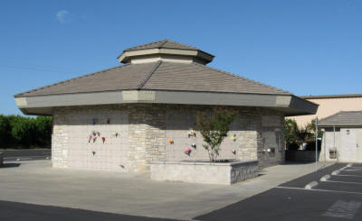 A small, round building with a stone facade and a peaked roof, featuring climbing holds on the walls and surrounded by a parking lot.