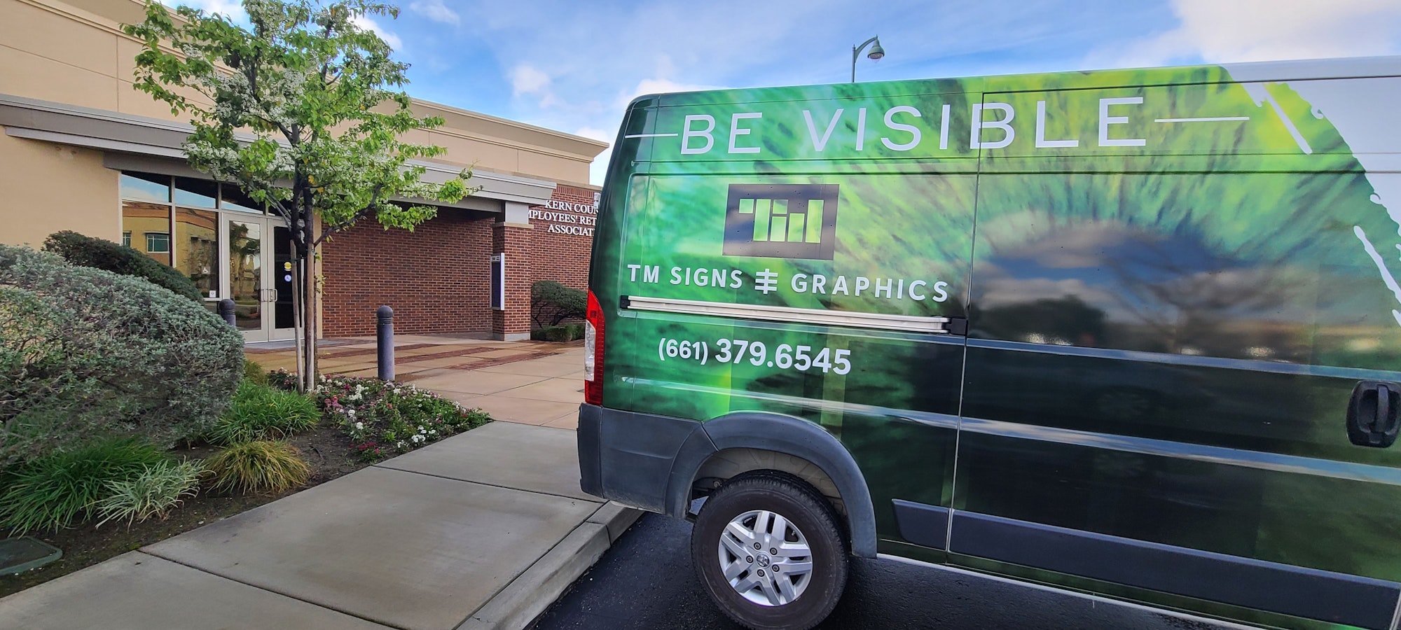 A van with "BE VISIBLE" and sign graphics advertisement parked near a building with "KERN COUNTY EYE CARE" sign.