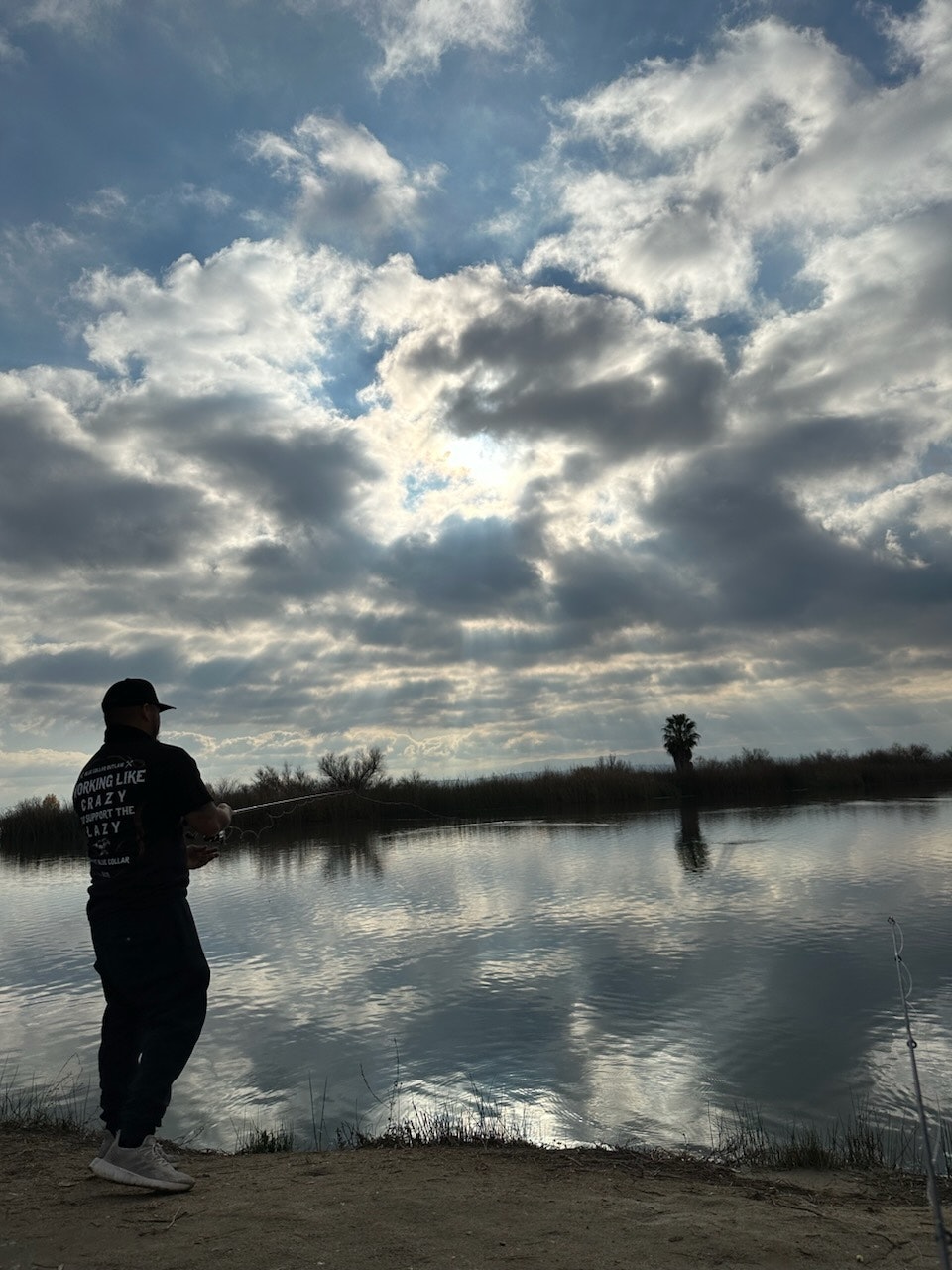 Person fishing by a calm lake with a dramatic cloudy sky overhead.