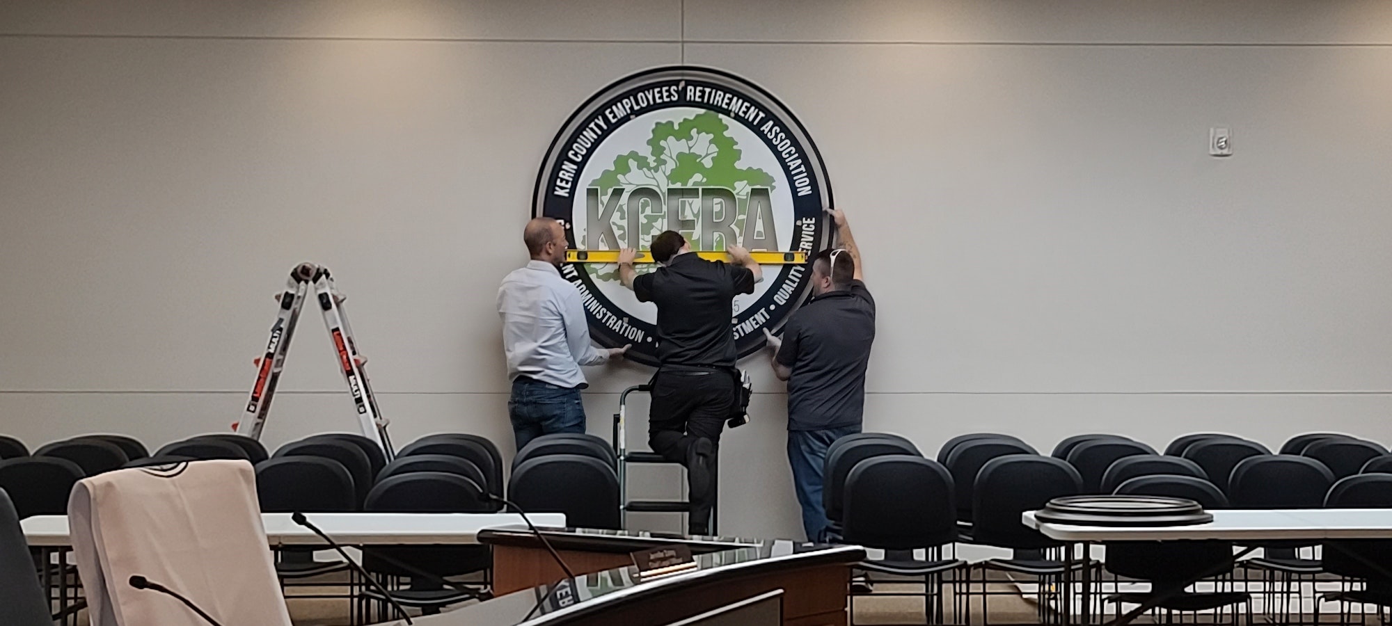 Three people are hanging a logo on a wall in a conference room with chairs and a speaker podium.