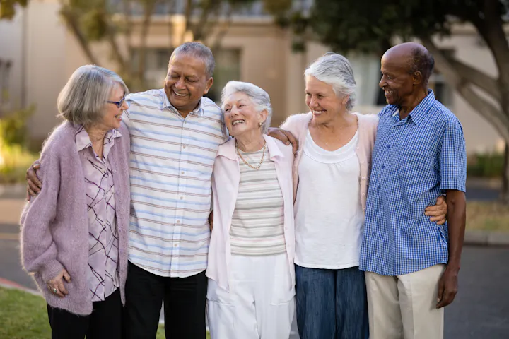 Five elderly people are happily standing together, arm in arm, outside.