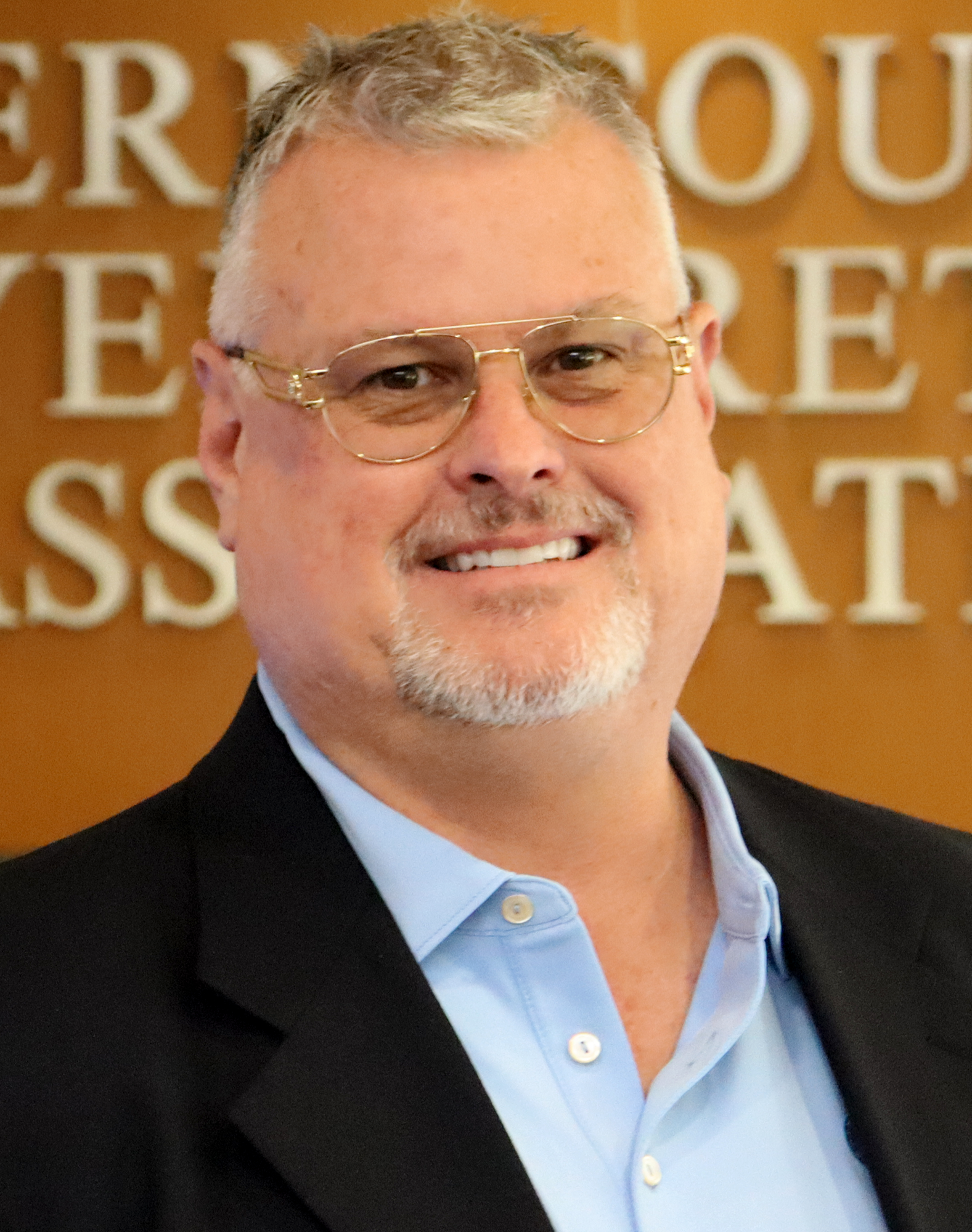 A man with glasses wearing a suit and blue shirt smiles in front of a blurred background with text.
