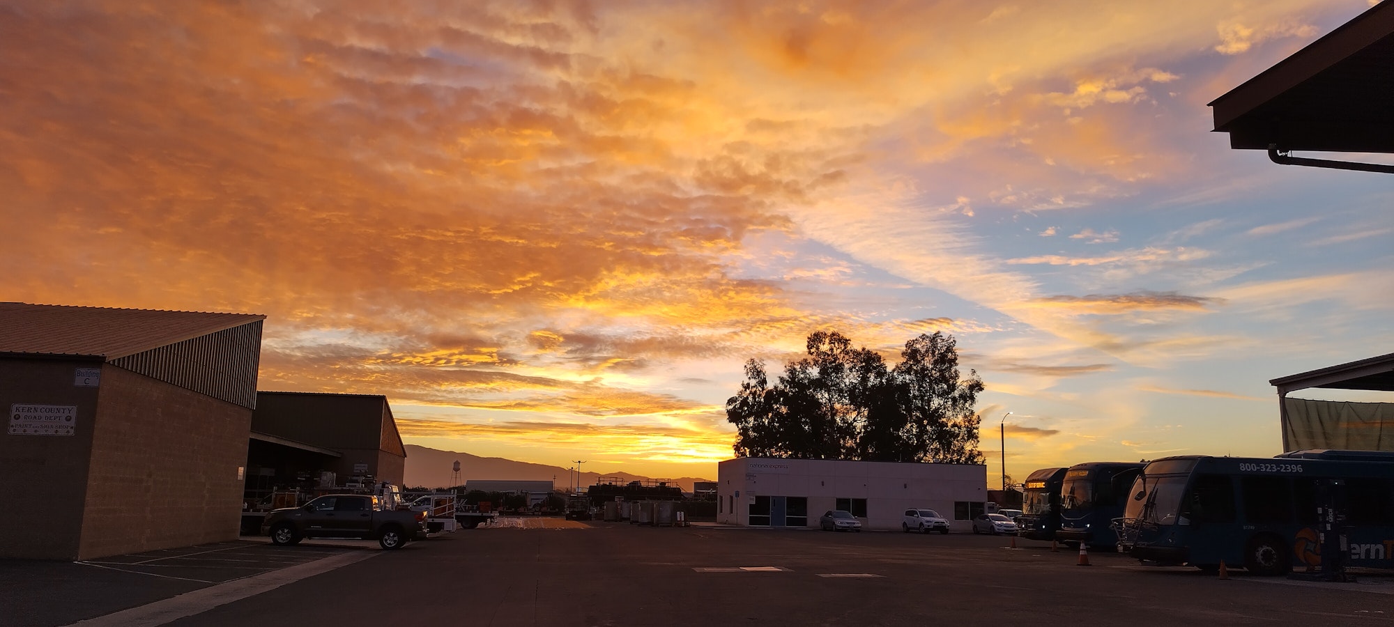 A vibrant sunset with orange clouds over industrial buildings, buses parked, and distant mountains.