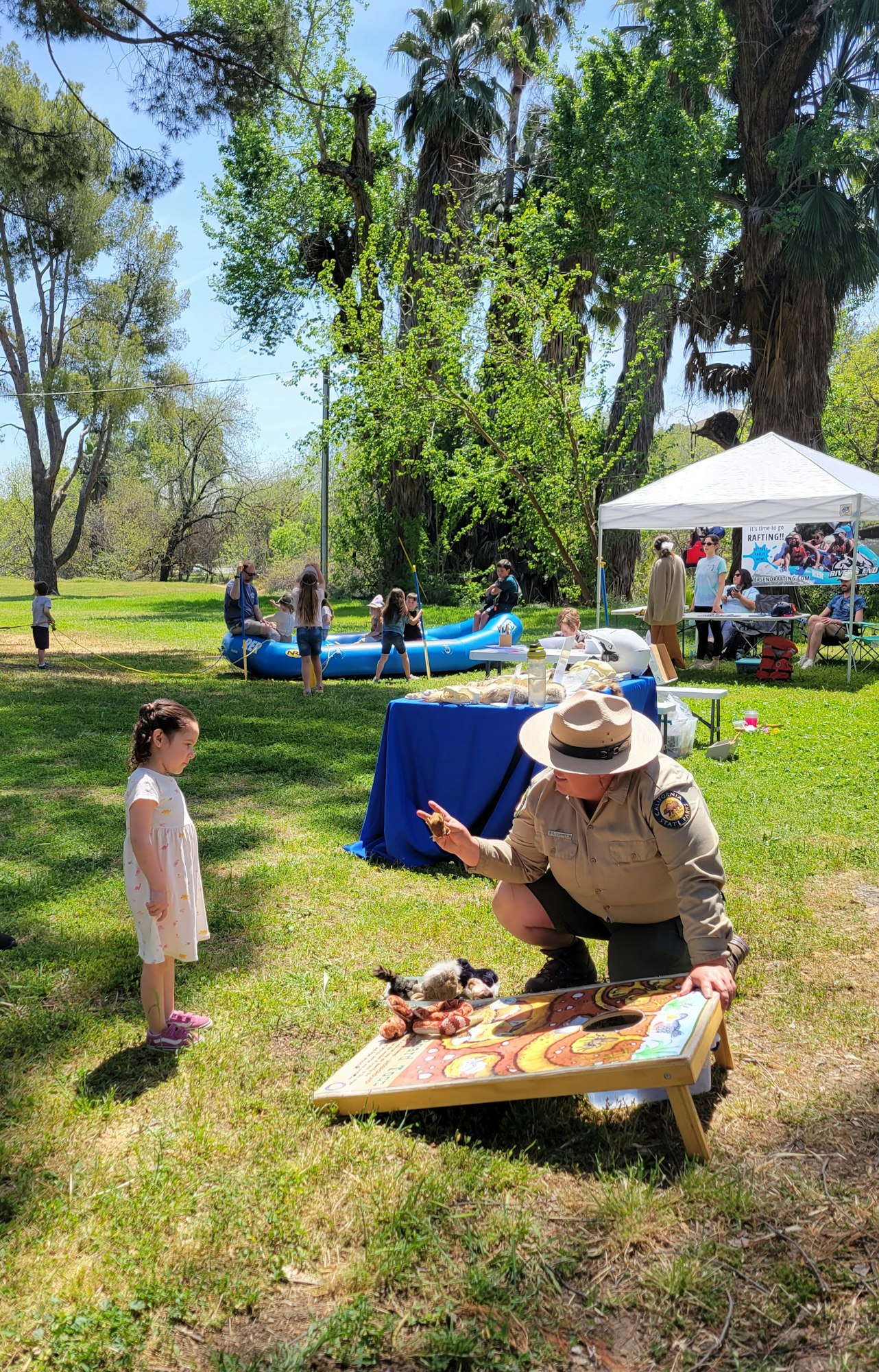Outdoor event with a ranger teaching a child, people in background, green trees, and blue sky.