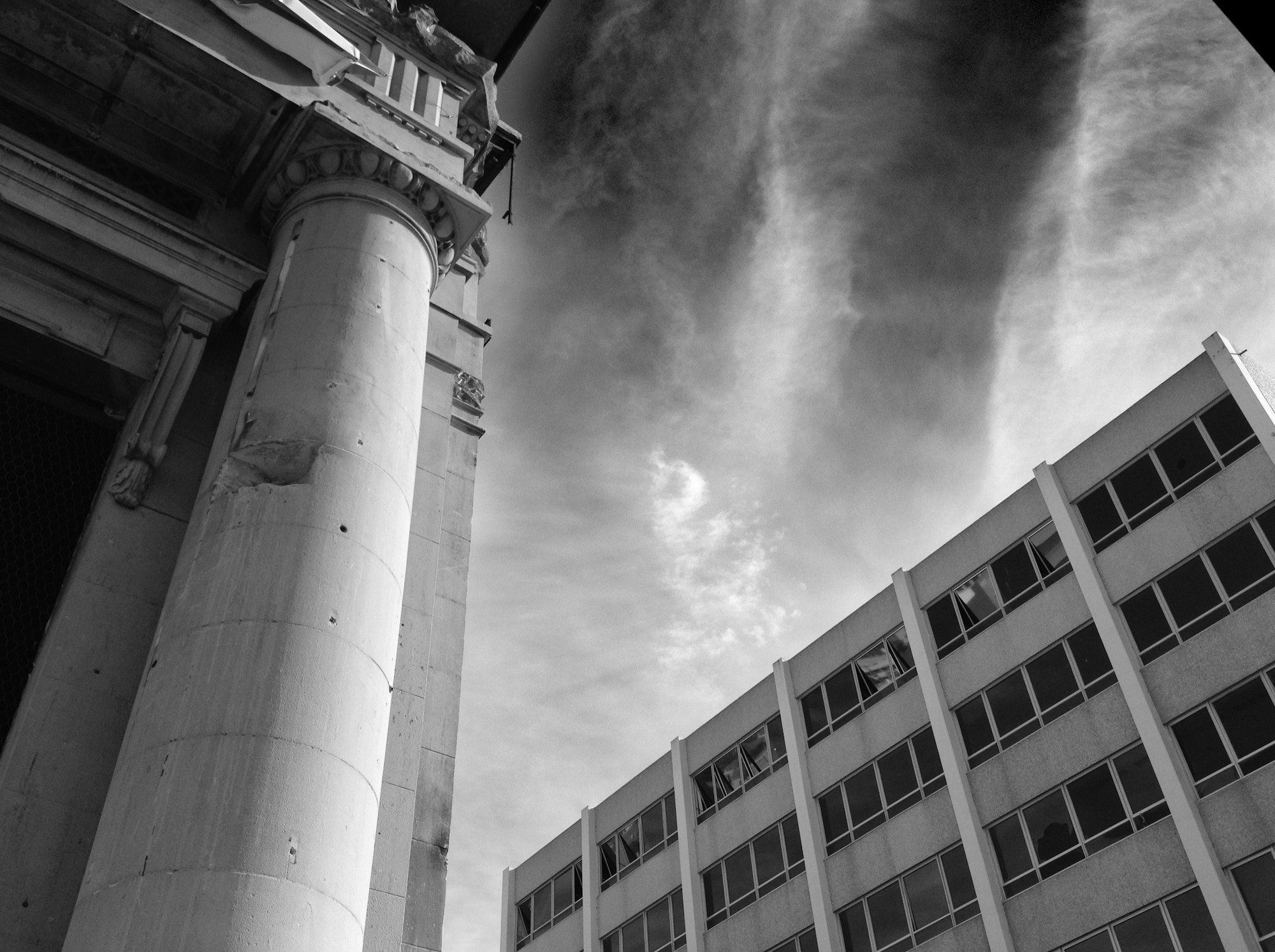 Black and white photo of a classical column and modern building against a cloudy sky.