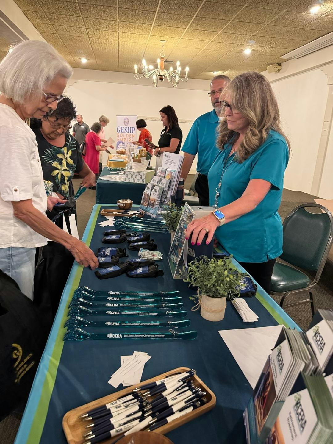 A busy event with people engaging at a table displaying promotional items like pens and brochures.