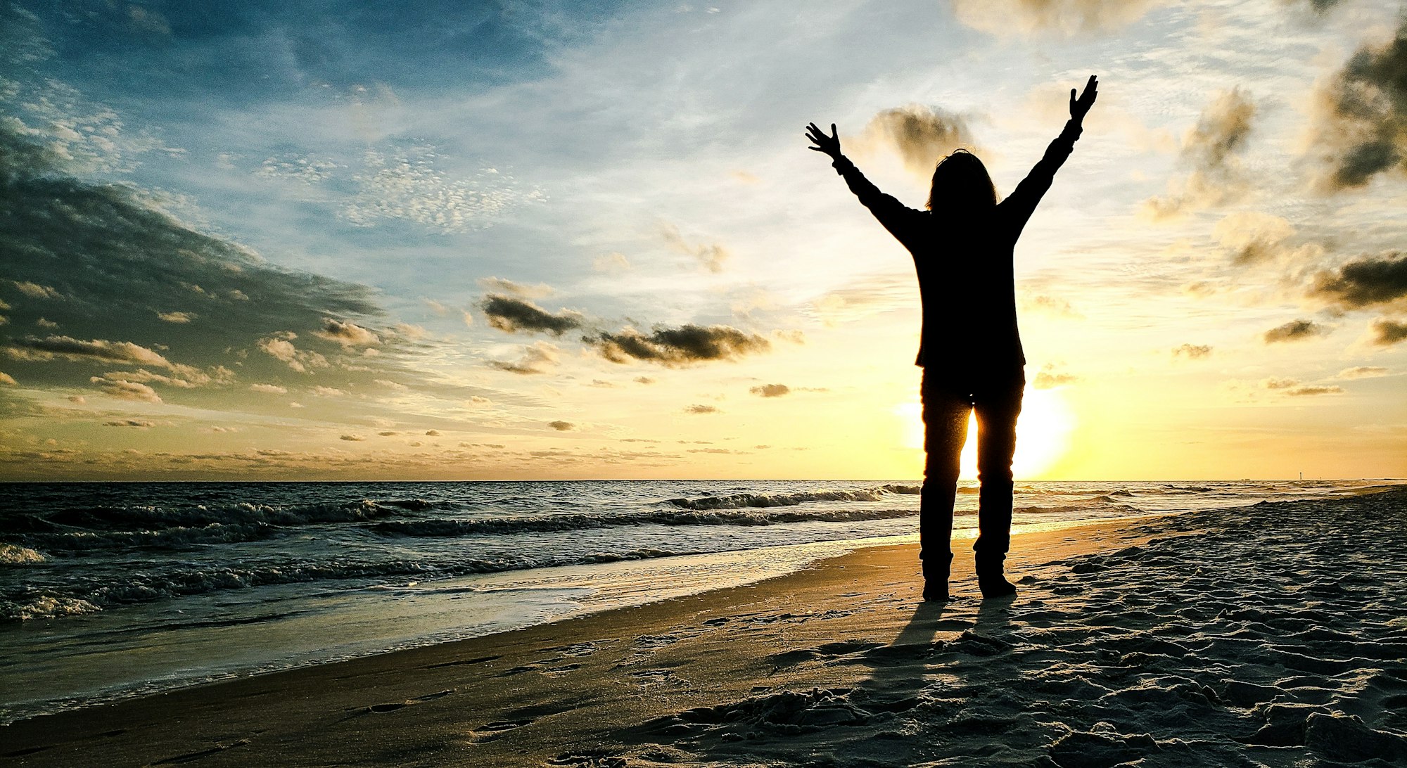 A silhouette of a person with arms raised, standing on a beach at sunset. Waves gently lap at the shore under a colorful sky.